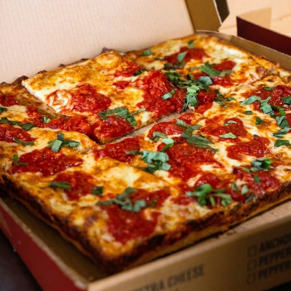 Square-cut Detroit-style pizza with rich red sauce, melted cheese, and fresh basil in a pizza box.