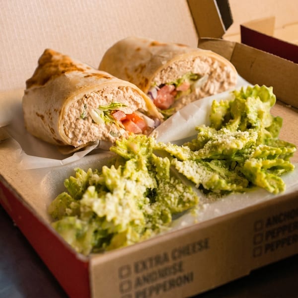 Two tuna salad wraps with lettuce and tomato, alongside green pasta salad, in a takeout box.